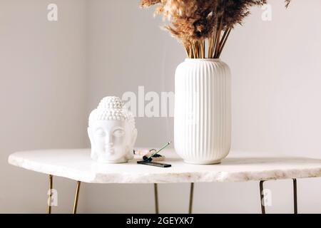 Small marble table with decorations, white vase with dry flowers, little buddha head statue and burning incense stick on stand in modern simplistic ap Stock Photo