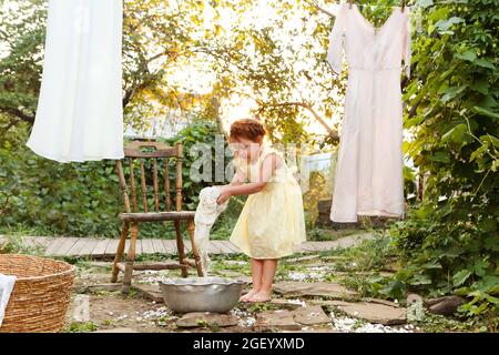 Cute little girl in dress washing white clothes in metal basin in backyard, child helping with chores, hanging laundry on clothesline and leaving it t Stock Photo