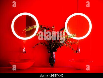 Photo of modern bathroom in red tones, two round shapes stylish wash basins, transparent vase with branches and leaves, pair of small size round illum Stock Photo