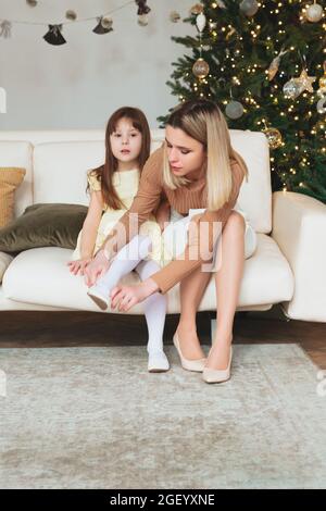 Photo of young mother and child dressed for New Years eve celebration, loving mom helping little girl put on shoes while sitting on sofa in living roo Stock Photo