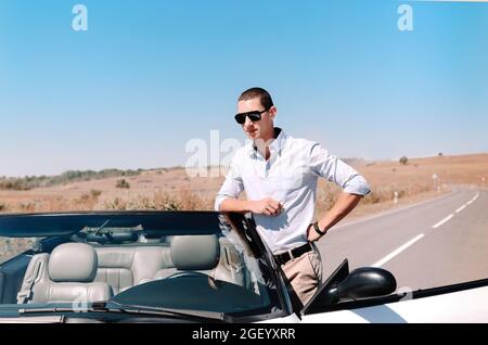 Handsome young man in black sunglasses leaning on luxury white convertible, successful businessman wearing casual clothes parked in middle of road in Stock Photo