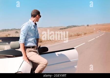 Handsome young man in black sunglasses leaning on luxury white convertible, successful businessman wearing casual clothes parked in middle of road in Stock Photo