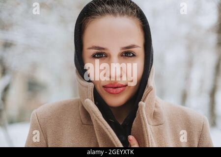 Young positive woman with funny smile on her face stands outside in beige coat and black scarf on head, shivering from winter cold weather against bac Stock Photo