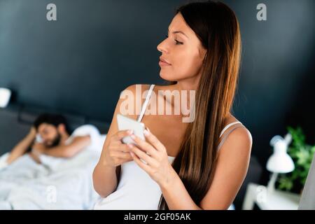 Young woman using cellphone while her husband sleeping on bed Stock Photo