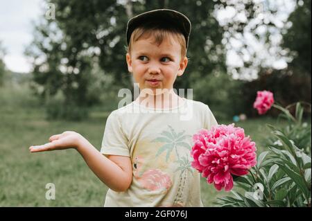 Russia, June 2020 - portrait of a cute touching little four year old kid boy with a pink peony flower in full bloom in the village garden in the count Stock Photo