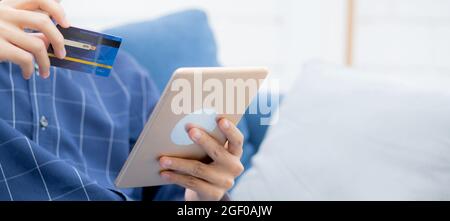 Young asian man smiling holding credit card shopping online with tablet computer buying and payment, male using debit card purchase or transaction of Stock Photo