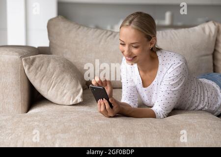 Happy blonde millennial smartphone user relaxing on soft couch Stock Photo