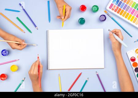 Kid hands holding pencils brushes to paint or draw in blank art sketchbook notebook. Top view to creative art work supplies on gray desk. Back to Stock Photo