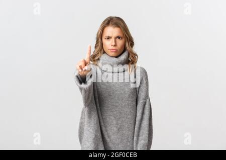 Image of serious and angry blond woman telling to stop, shaking finger disappointed, scolding someone for doing bad thing, standing over white Stock Photo