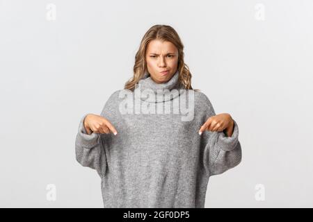 Image of doubtful and skeptical blond girl, frowning and pouting displeased, pointing fingers down at something bad, standing over white background Stock Photo