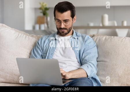 Serious millennial man in glasses using laptop, relaxing on couch Stock Photo