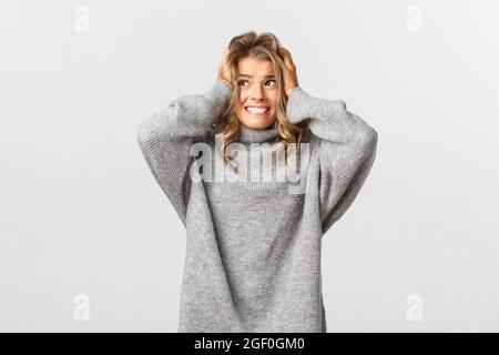 Portrait of blond girl in panic, looking left and holding hands on head, feeling alarmed and distressed, standing over white background Stock Photo