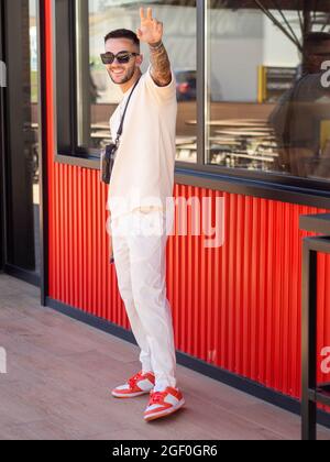 A young cheerful Spanish man with tattoos standing near a building and waving on a sunny day Stock Photo
