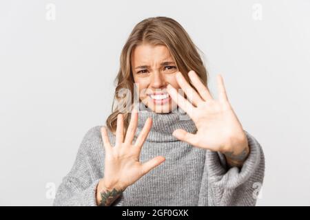 Close-up of bothered blond girl defending herself, grimacing from something disgusting or too bright, standing over white background Stock Photo