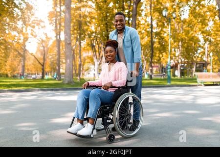 Full length of cheerful black guy taking his impaired girlfriend in wheelchair for walk at city park in autumn Stock Photo