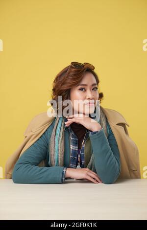 Attractive young woman wearing coat over warm clothes when sitting at table against yellow background Stock Photo
