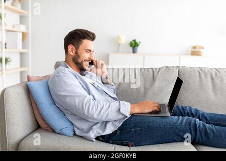 Home office concept. Millennial businessman talking on smartphone and working on laptop, sitting on couch Stock Photo