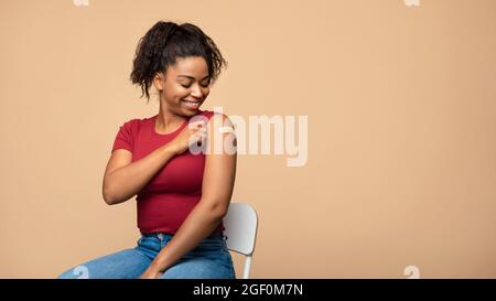 Cheerful vaccinated Afro woman showing plaster on her shoulder after immunization against covid over beige background Stock Photo