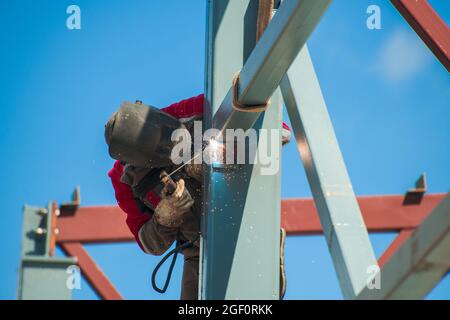 the welder performs welding work at a height while standing on a metal structure against the background of a blue cloudless sky Stock Photo