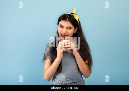 Lovely Indian teenage girl eating tasty chocolate bar, surrendering to temptation on blue studio background Stock Photo
