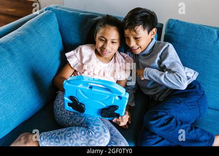 Happy latin siblings sitting on sofa using digital device at home in Mexico Stock Photo