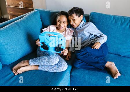 Happy latin siblings sitting on sofa using digital device at home in Mexico Stock Photo