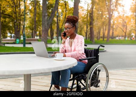 Disabled black lady in wheelchair using laptop, having conversation on smartphone at outdoor cafe in autumn park Stock Photo