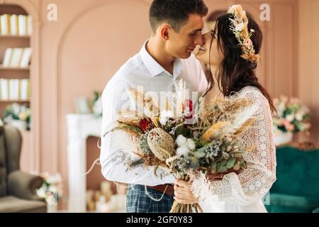Happy young couple, stylish groom in a suit and a young bride in a wedding lace dress with a bouquet of flowers, sit together, hug, in the studio on t Stock Photo