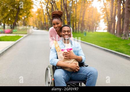 Cheerful handicapped man in wheelchair holding paper bag with food, his lovely girlfriend helping him in autumn park Stock Photo