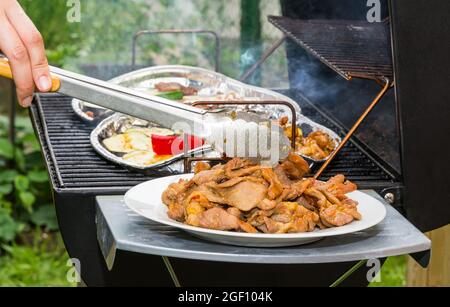 Grilling of hot pork meat and vegetable slices on charcoal grill on garden bbq party. Detail of human hand holding metal tongs. Barbecue meal on plate. Stock Photo
