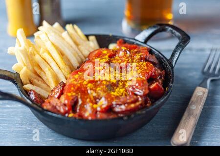 Currywurst with french fries in black pan served with beer on wooden table. Stock Photo