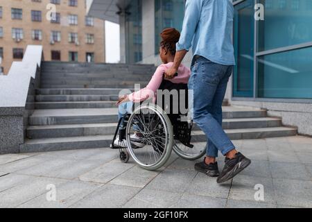 Black guy pushing wheelchair with handicapped woman, having problem entering building, standing near stairs without ramp Stock Photo