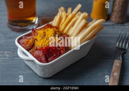 Currywurst with french fries in white bowl on wooden table served with beer. Stock Photo
