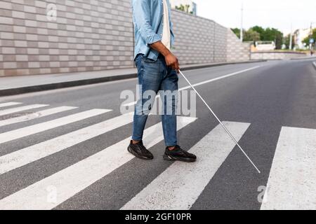 Unrecognizable young black visually impaired man with cane walking across city street, closeup Stock Photo