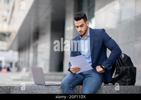 Arab entrepreneur sitting by office building with laptop, reading documents Stock Photo