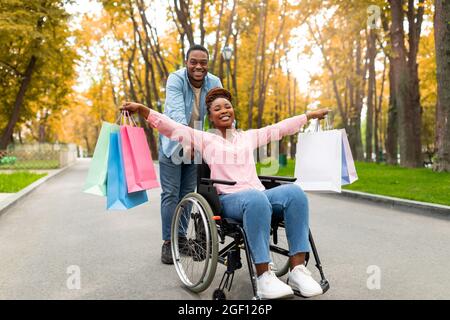 Young positive black guy pushing wheelchair with his disabled wife holding shopping bags at autumn park Stock Photo