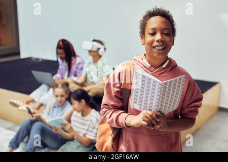 Waist up portrait of young schoolboy holding book and smiling at camera while standing in school hall, copy space Stock Photo