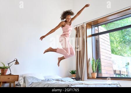 Happy carefree young African American woman wearing pajamas jumping on bed. Stock Photo