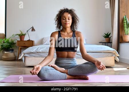Young calm African woman wearing sportswear sitting at home bedroom doing yoga. Stock Photo