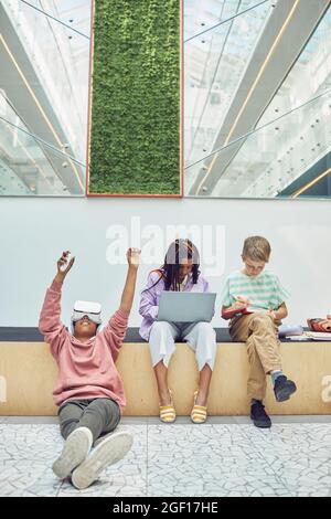 Vertical full length portrait of contemporary children using devices in modern school, focus on boy wearing VR in foreground Stock Photo