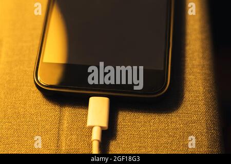 detail of a smart mobile phone charging its battery by a cable placed on the bedside table of the bedroom disturbing the sleep of the owner concept da Stock Photo