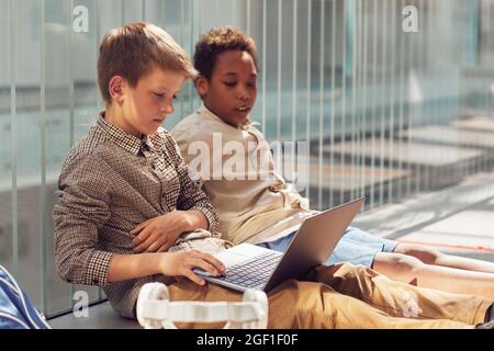 Portrait of two schoolboys using laptop while sitting on floor in school lit by sunlight, copy space Stock Photo