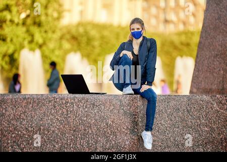 Woman in facial mask with laptop sits outdoors. Stock Photo