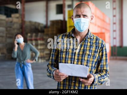 Male manager in protective mask with documents inside warehouse Stock Photo