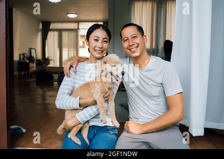 Young adult Asian couple holding a puppy with home interior in background. 30s mature woman holding a dog pet talking with a man. Husband and wife living together at house. Family happiness concept Stock Photo