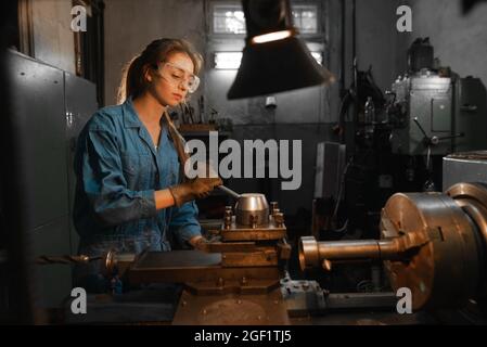 A young woman in goggles and overalls is standing near the lathe. Profession concept Turner, Metalworking, Turning, Industry, Metal. Stock Photo