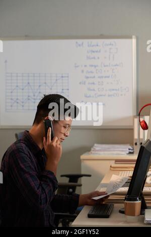 Young programmer looking at document with project flowchart and talking on phone with colleagues from marketing department Stock Photo