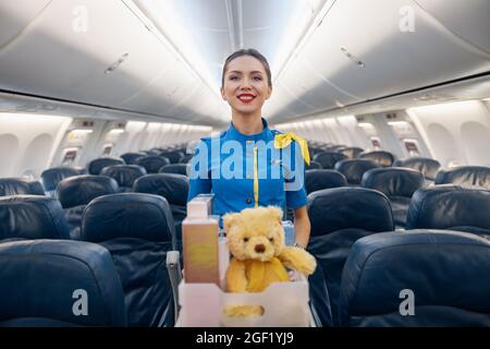 Attractive female air hostess in bright blue uniform smiling at camera while leading trolley cart with gifts, souvenirs through empty plane aisle Stock Photo