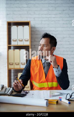Pensive frowning construction engineer sitting at office desk with blueprints and looking away though window Stock Photo