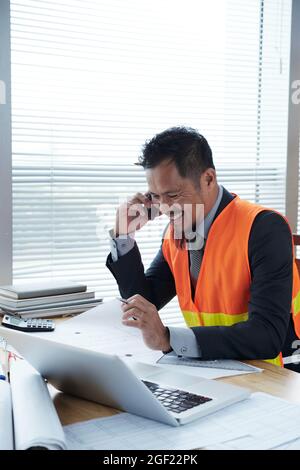 Positive construction engineer working at office desk, checking blueprints and discussing readiness of construction site with contractor on phone Stock Photo